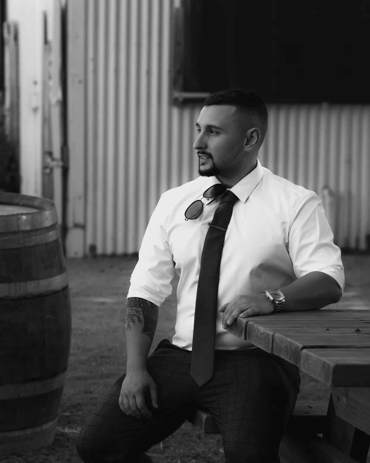 Nik of Royale Photography in a white shirt and tie, seated at an outdoor table beside a wine barrel, photographed in black and white.
