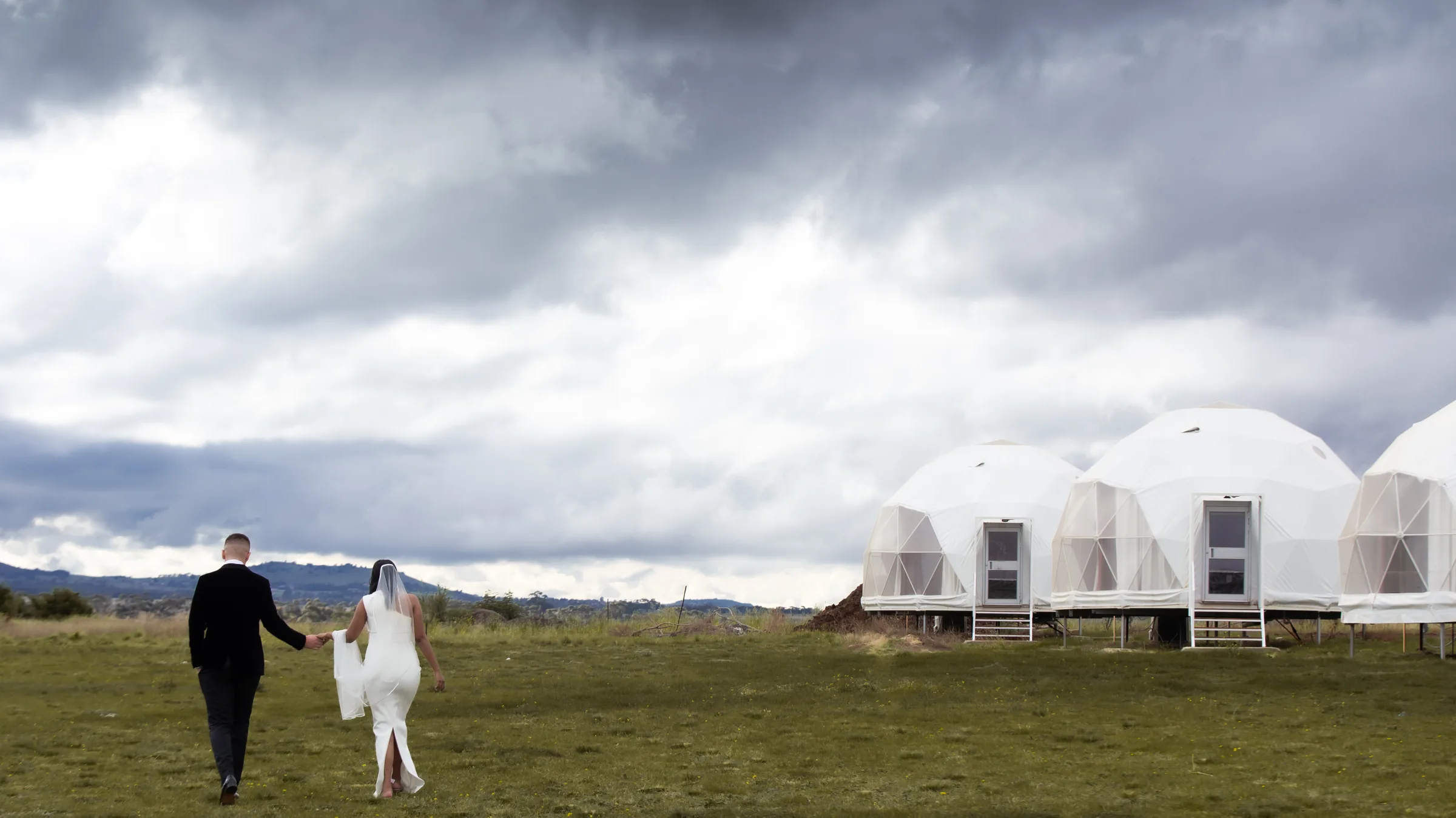 A bride and groom walk hand in hand across an open field toward geodesic dome tents beneath a dramatic storm sky, photographed by Royale Photography in regional Victoria.