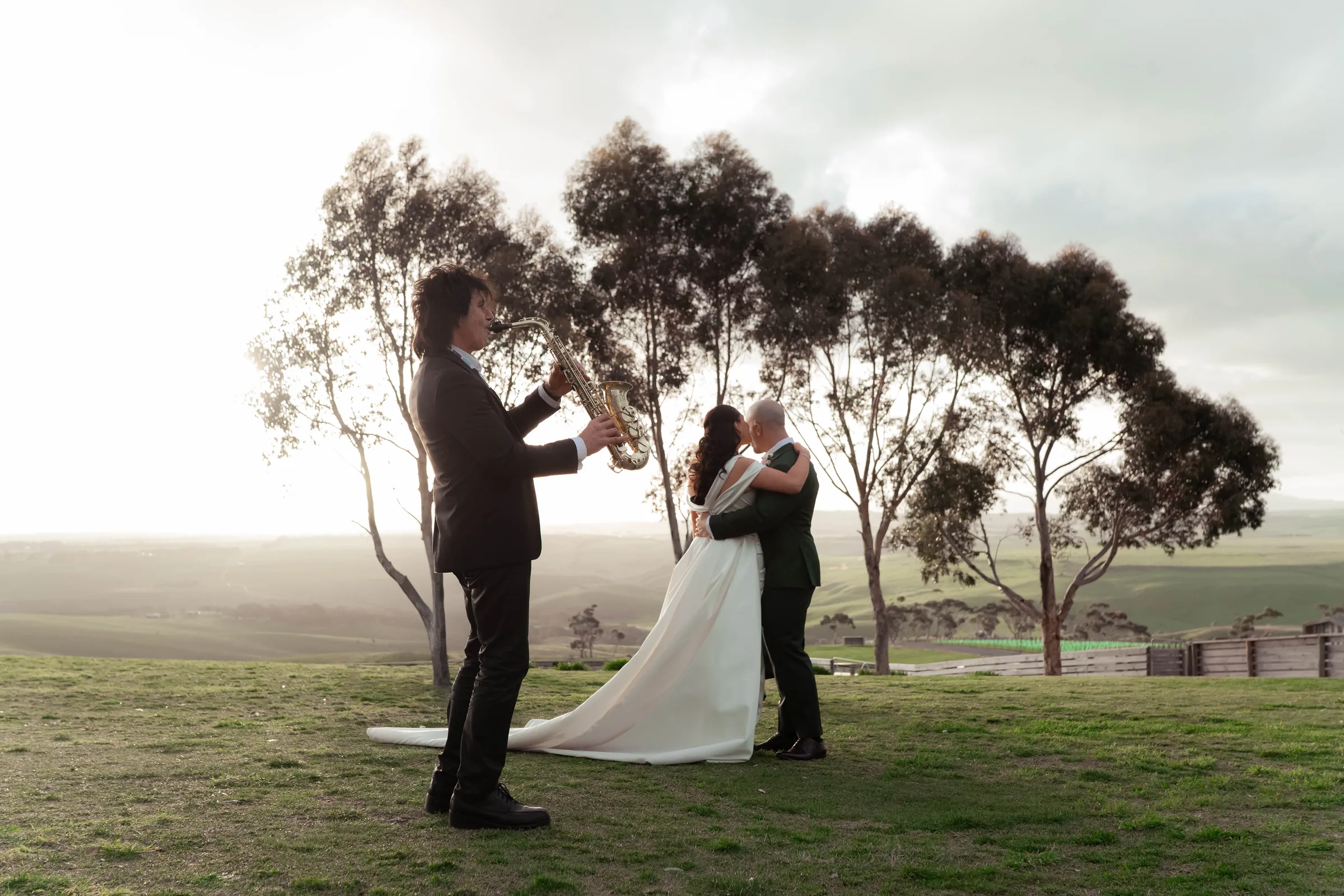 A couple embrace at a hilltop ceremony at dusk while a saxophonist plays, photographed by Royale Photography.