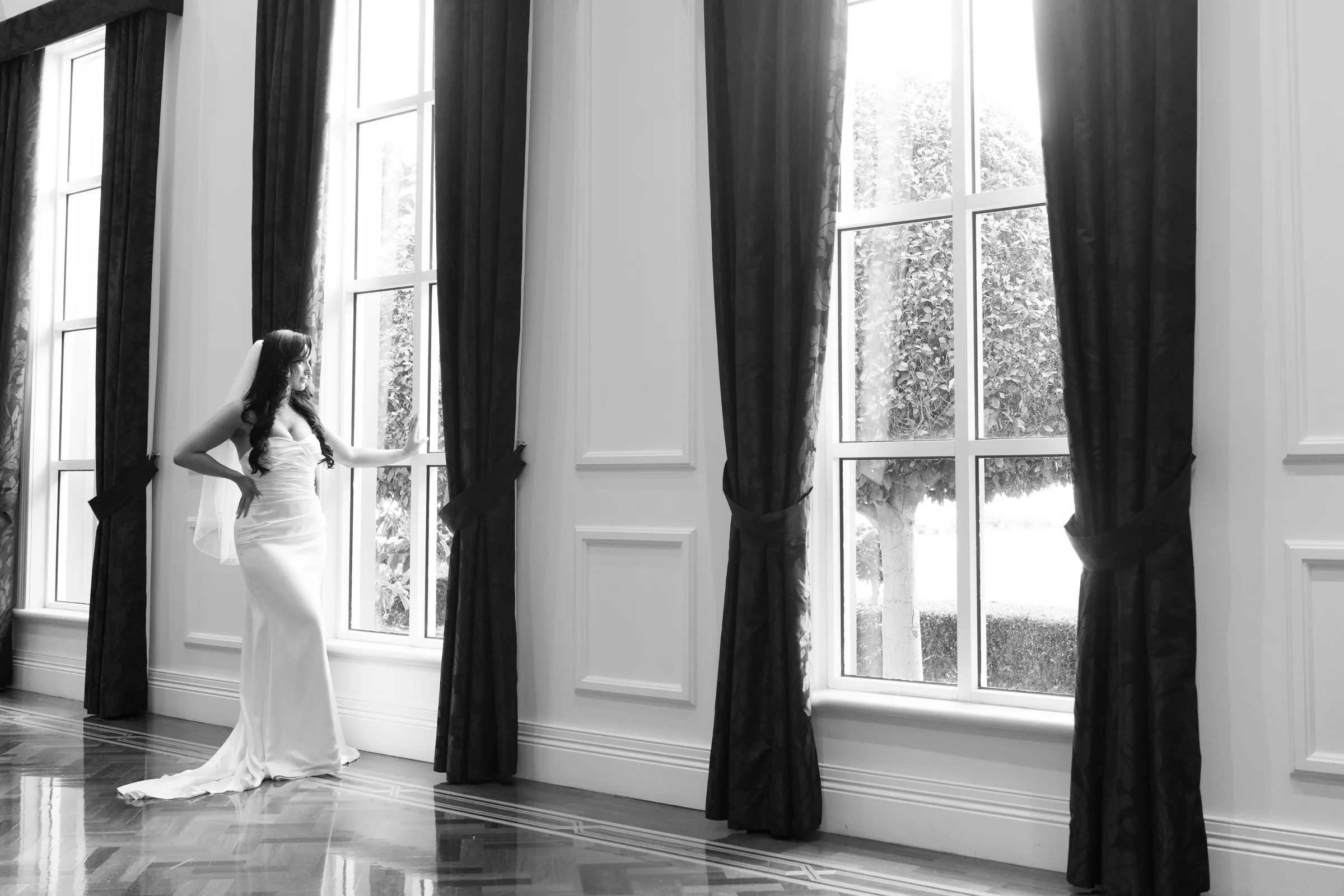 Black and white portrait of a bride beside tall windows in an elegant hall, photographed by Royale Photography.